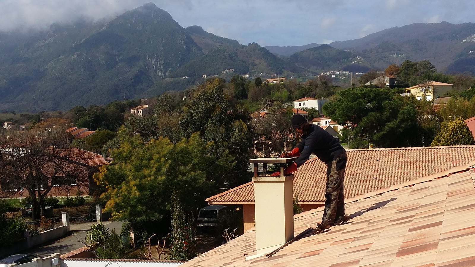 Ramoneur penché sur une souche de cheminée pour inspecter le conduit, montagnes corses en fond