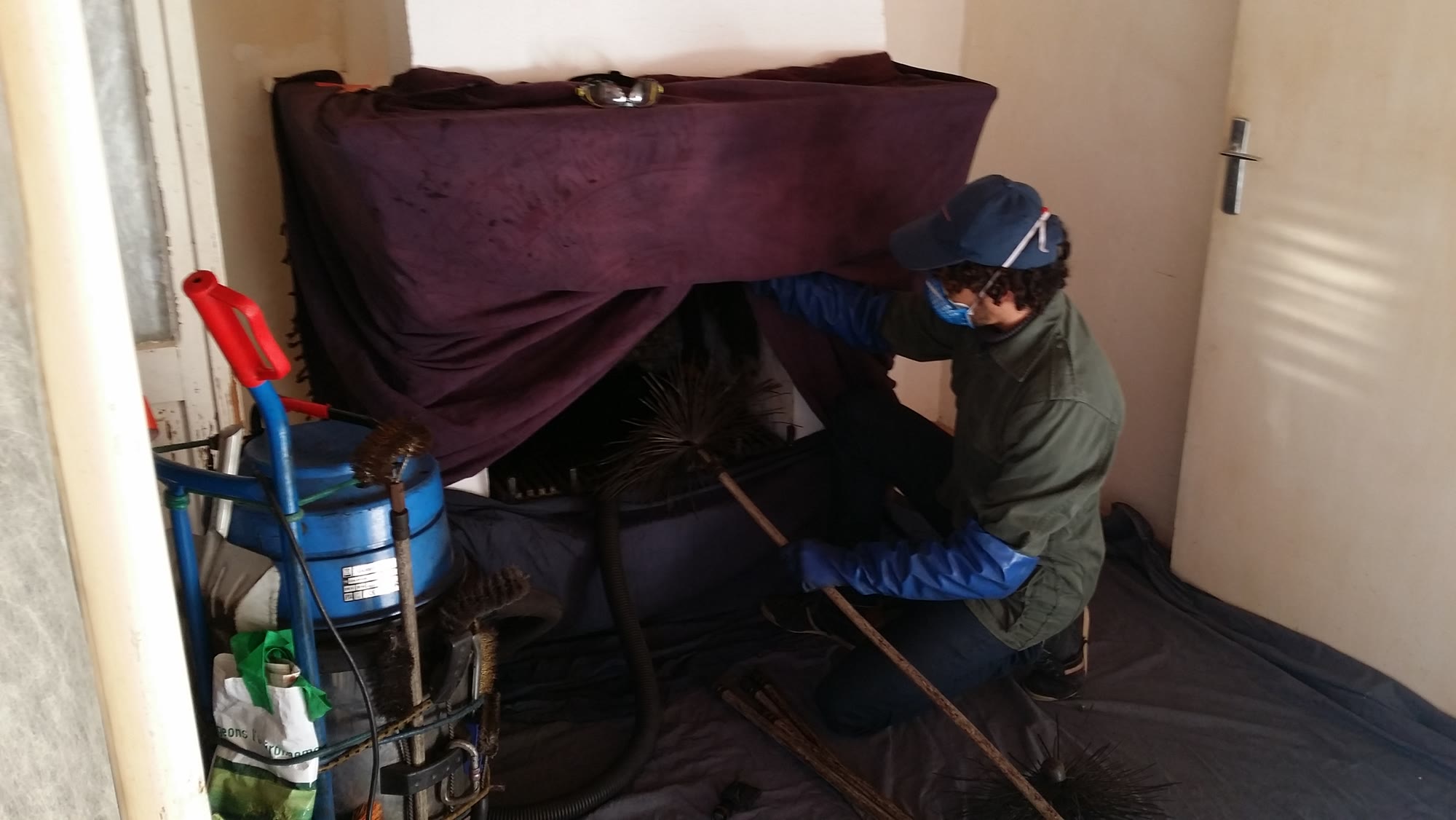 Ramoneur agenouillé devant un foyer, mobilier protégé par une bâche, aspiration en cours